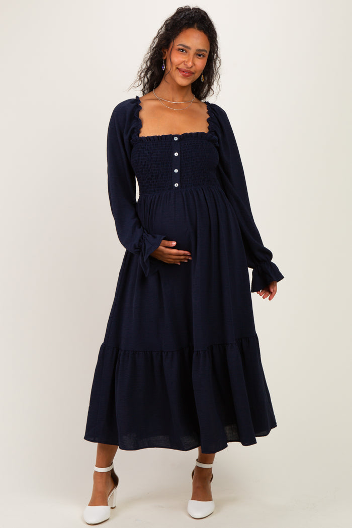 woman wearing a navy blue smocked maternity dress with long sleeves and tiered hem, showcasing her baby bump