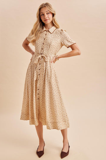 woman wearing a beige floral midi dress with short sleeves and a tie waist detail, posing with one hand on her hip
