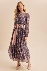 woman wearing a flowy purple floral maxi dress with long sleeves and a pink tie belt