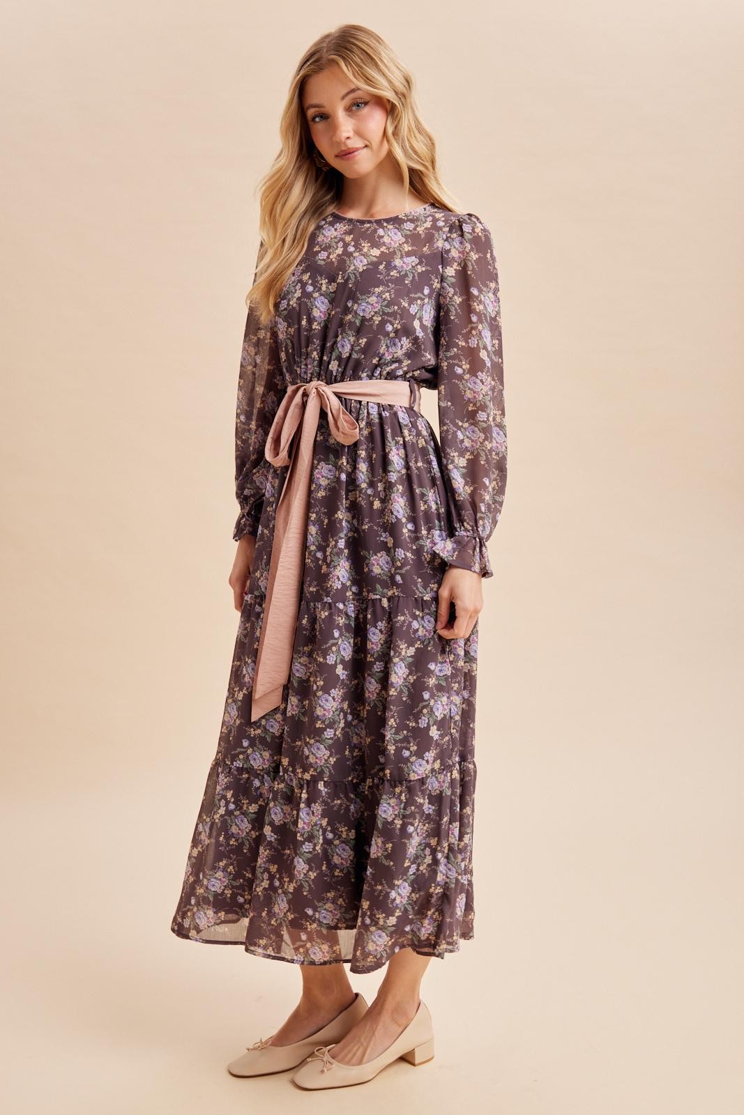 woman wearing a long floral maxi dress with long sleeves and a belt, standing against a neutral background