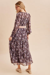 woman wearing a dark floral maxi dress with long sleeves and a tiered skirt, showing a pink belt at the waist from the back