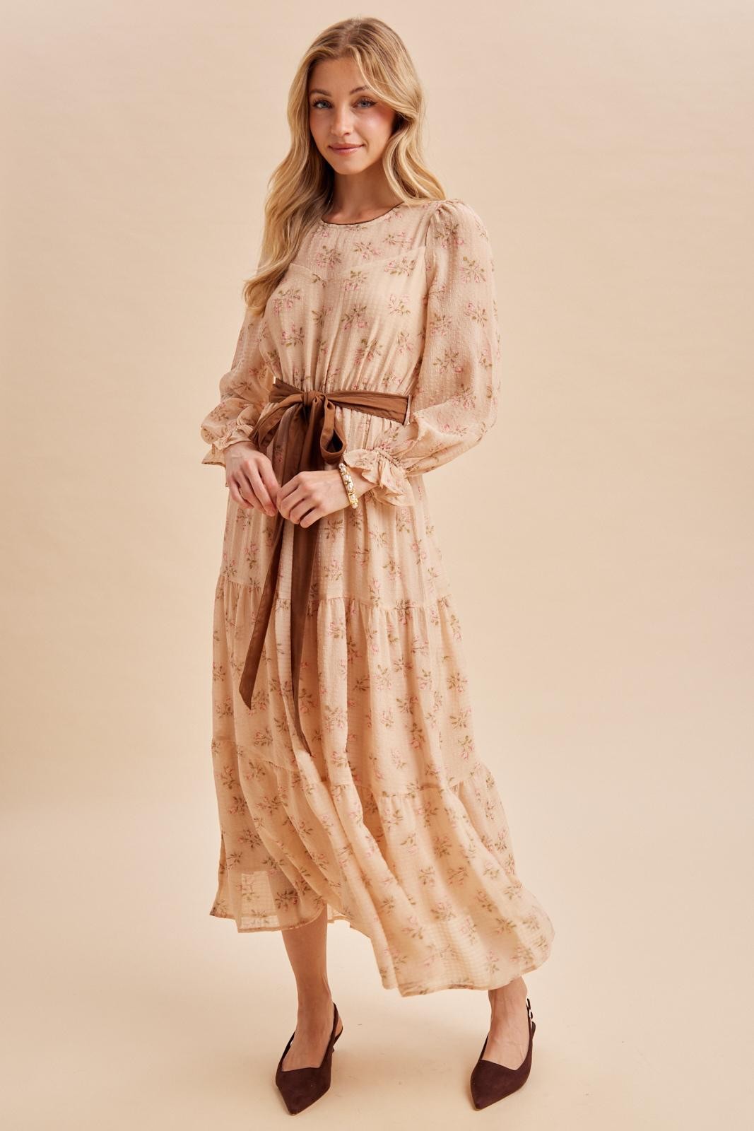 woman wearing a floral long sleeve maxi dress with a tie belt and flared hem, posing against a neutral background