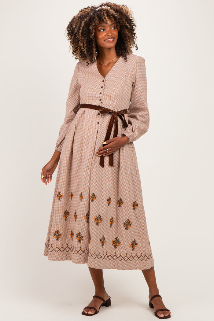 woman wearing a beige maternity dress with floral embroidery and long sleeves, styled with a brown belt