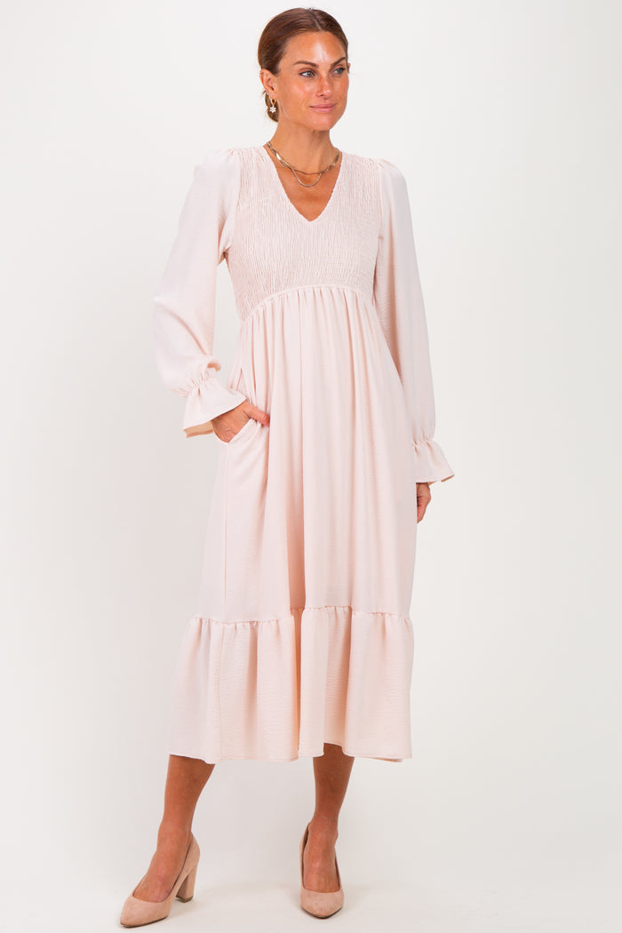 woman wearing a blush pink long sleeve dress with a smocked bodice and ruffle hem, standing with hands in pockets