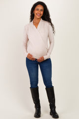 woman wearing a light pink wrap maternity top and dark blue jeans, with a visible baby bump, standing in tall black boots