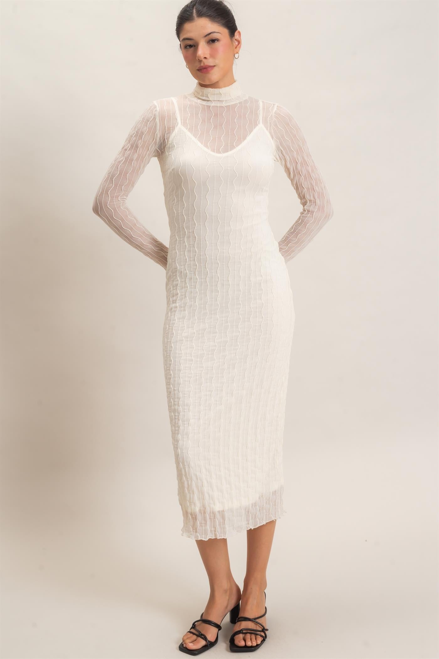 woman wearing a white ribbed midi dress with sheer long sleeves and a high neckline, styled with black strappy heels