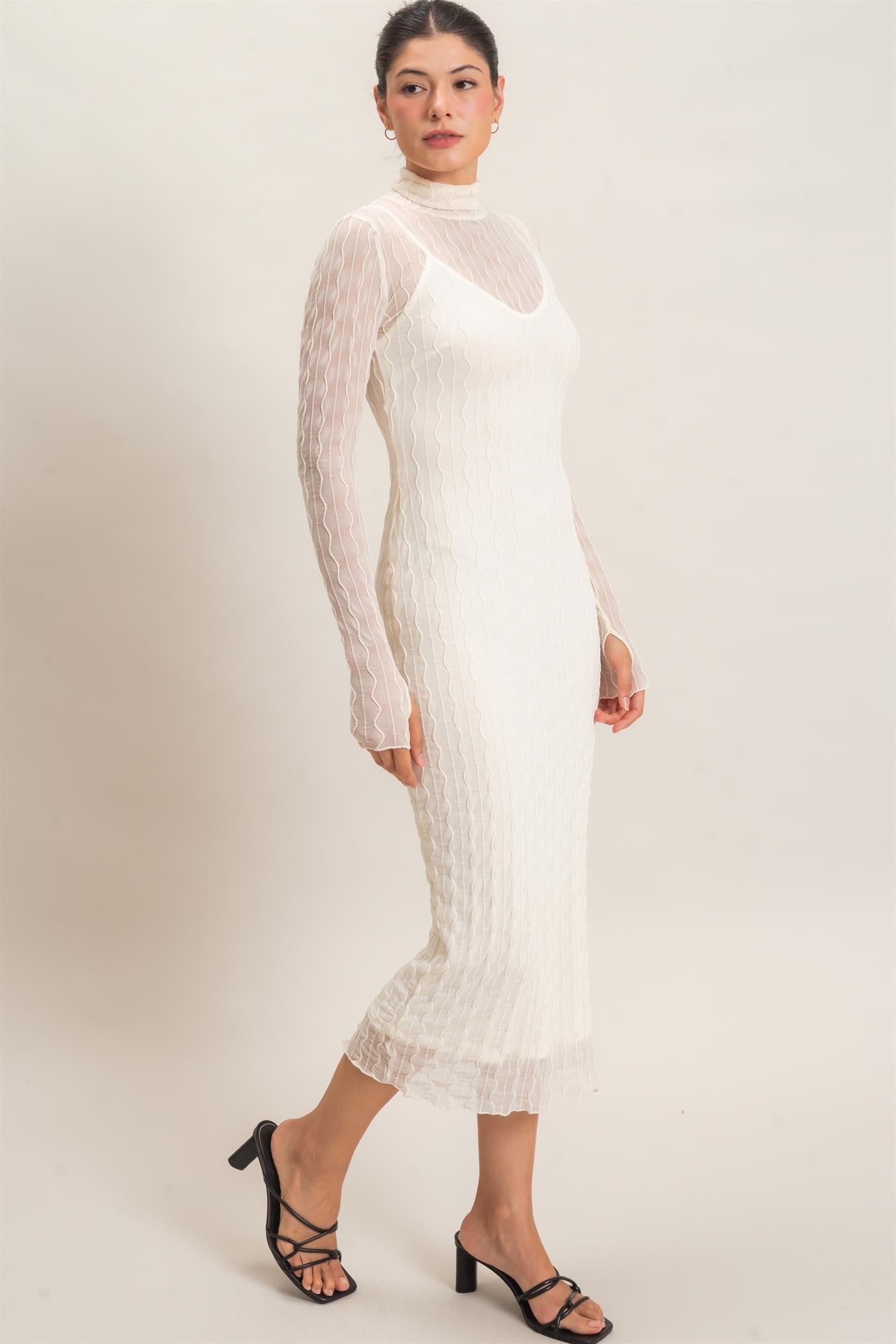 woman wearing a white fitted midi dress with sheer long sleeves and a scalloped hem, styled with black strappy heels