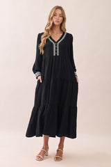 woman wearing a black tiered maxi dress with long sleeves and decorative detailing on the neckline and cuffs