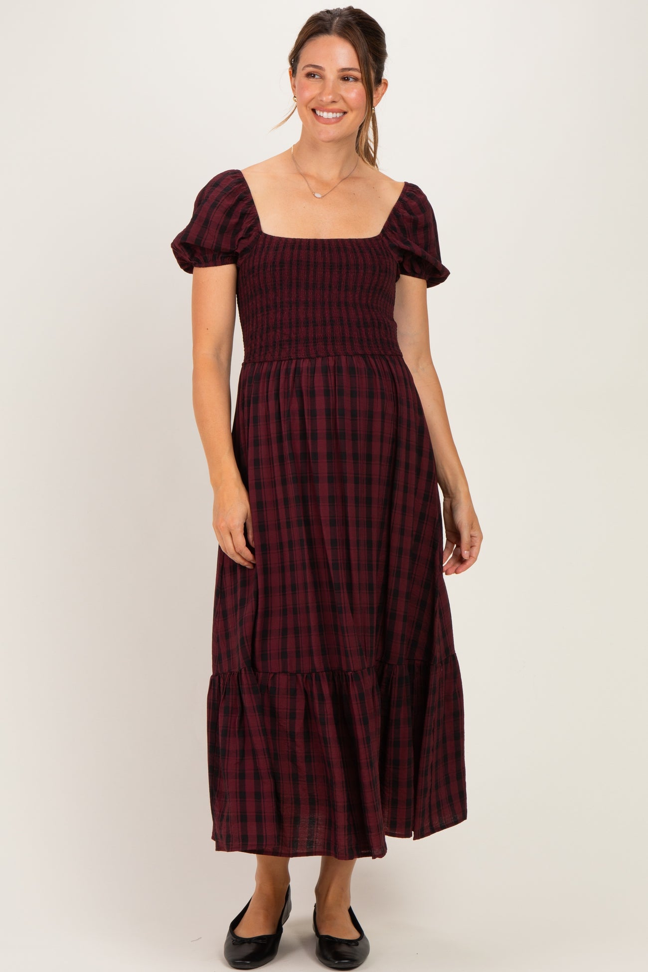 woman wearing a burgundy and black checked maternity dress with puff sleeves and a smocked bodice, standing and smiling