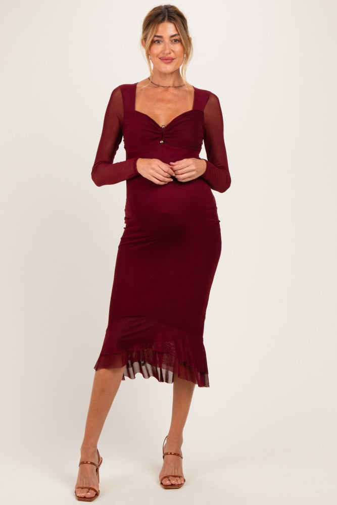 woman wearing a burgundy maternity dress with long sheer sleeves and a ruffled hem, showcasing her baby bump
