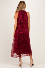 woman wearing a burgundy maternity dress with floral embroidery and a tie-back detail, showing a flowing length and a sleeveless design