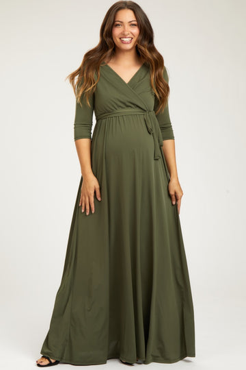 woman wearing a dark green maternity wrap maxi dress with three-quarter sleeves and a tied waist, standing and smiling