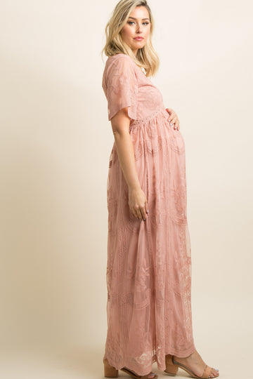 woman wearing a pink lace maternity gown with a floral bouquet and holding her baby bump while sitting in a grassy field