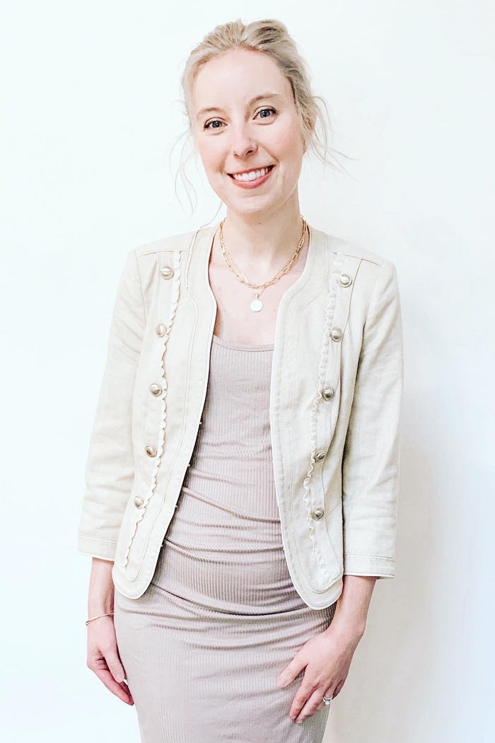 woman wearing a beige maternity jacket over a fitted beige dress, smiling with a neutral background