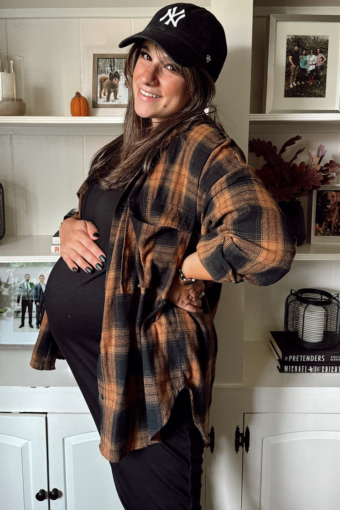 woman wearing a black maternity dress and an orange and black plaid shirt, showing her baby bump and smiling