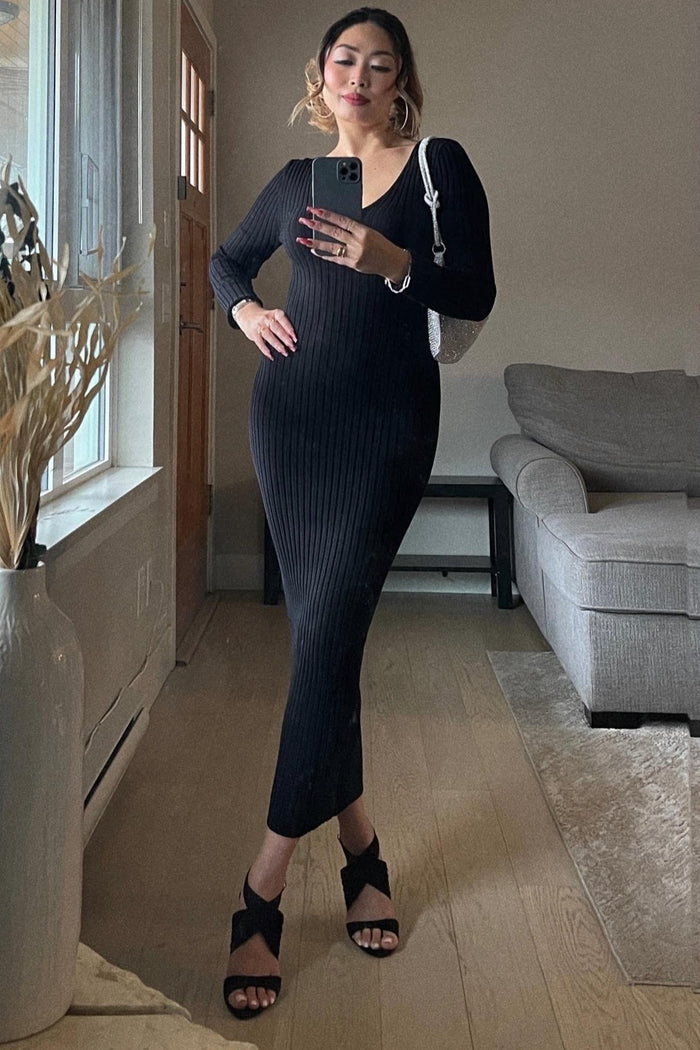 woman wearing a black ribbed long sleeve fitted dress and high heels, posing in a bright room with a plant