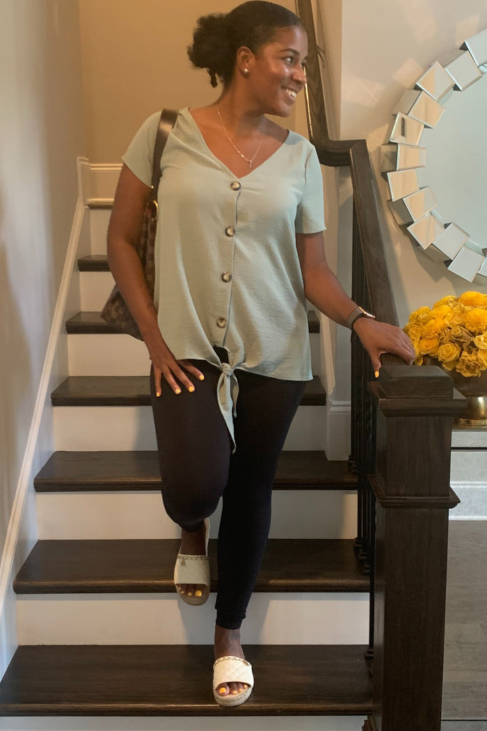 woman wearing a light green short sleeve top with buttons and a tie front, paired with black leggings and sandals, standing on stairs