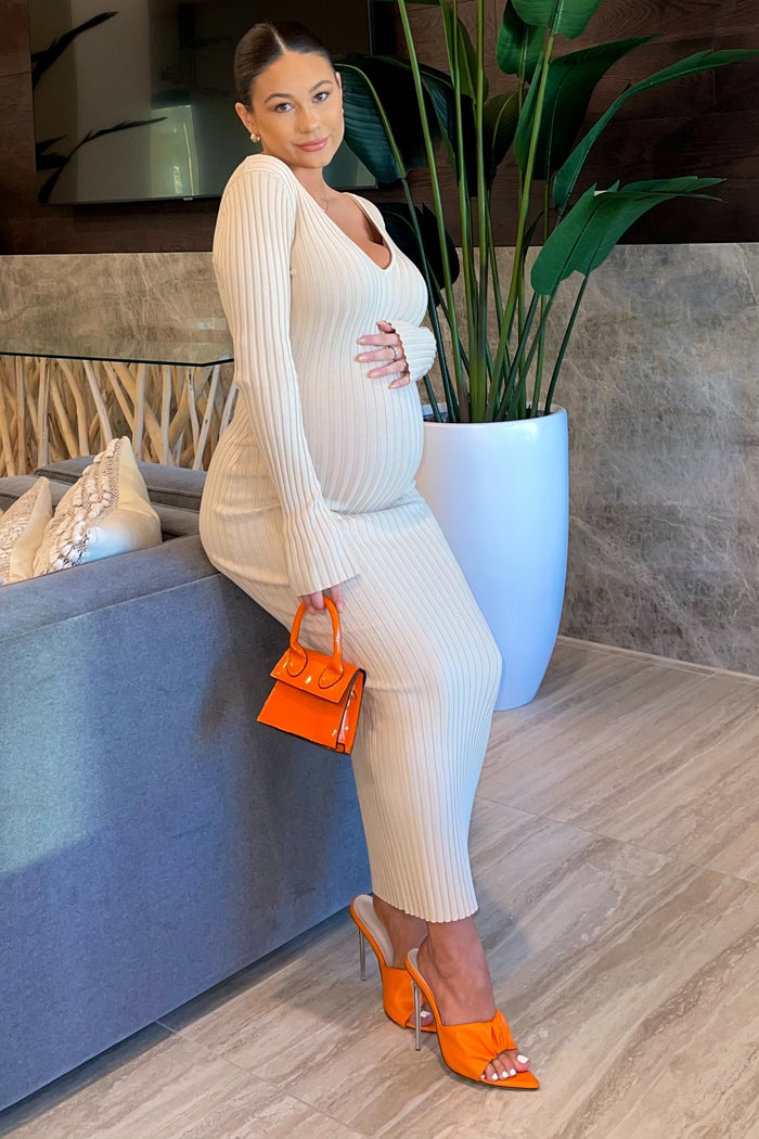 woman wearing a beige ribbed maternity dress, holding her baby bump, complemented by orange accessories and heels