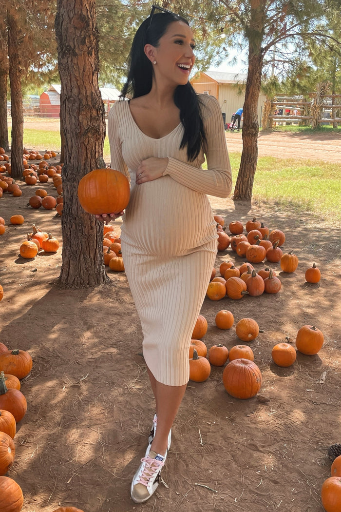 woman wearing a ribbed light beige maternity dress and holding a pumpkin, showing her baby bump among pumpkins in a field