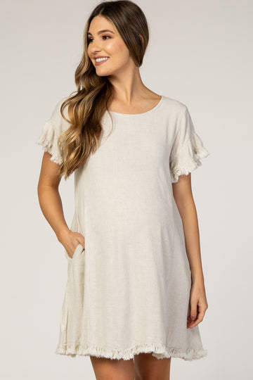 woman wearing a cream maternity dress with fringe detailing on the sleeves and a relaxed fit, smiling and holding her baby bump