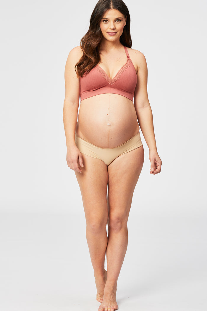 woman wearing a rust-colored lace bralette and nude maternity bikini underwear, showing her baby bump