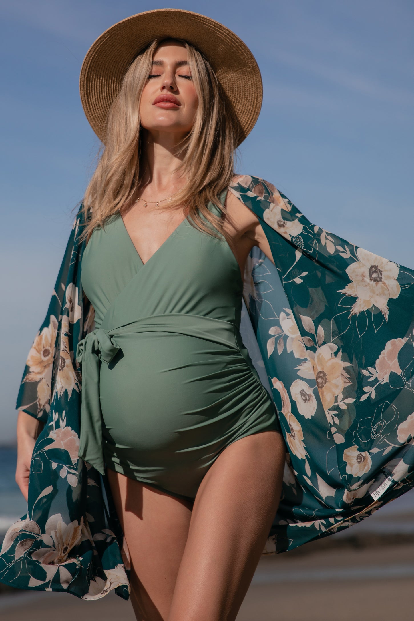 woman wearing a green maternity swimsuit with a floral kimono and a straw hat, showing her baby bump