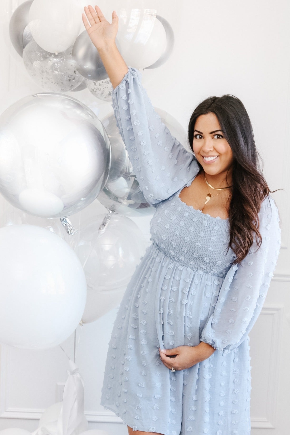 woman wearing a light blue maternity dress with textured details and holding her baby bump, standing near balloons