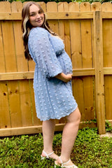 pregnant woman in light blue swiss dot dress posing in front of a wooden fence