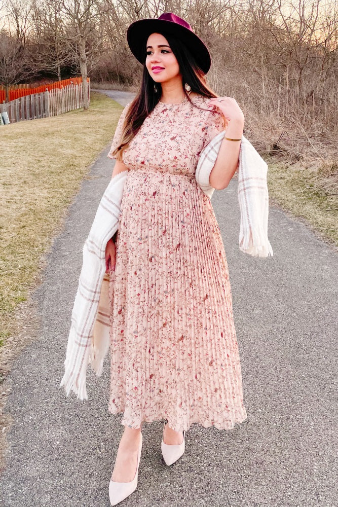 woman wearing a floral pink maxi dress with pleats and a wide-brim hat, holding a light shawl while walking outdoors