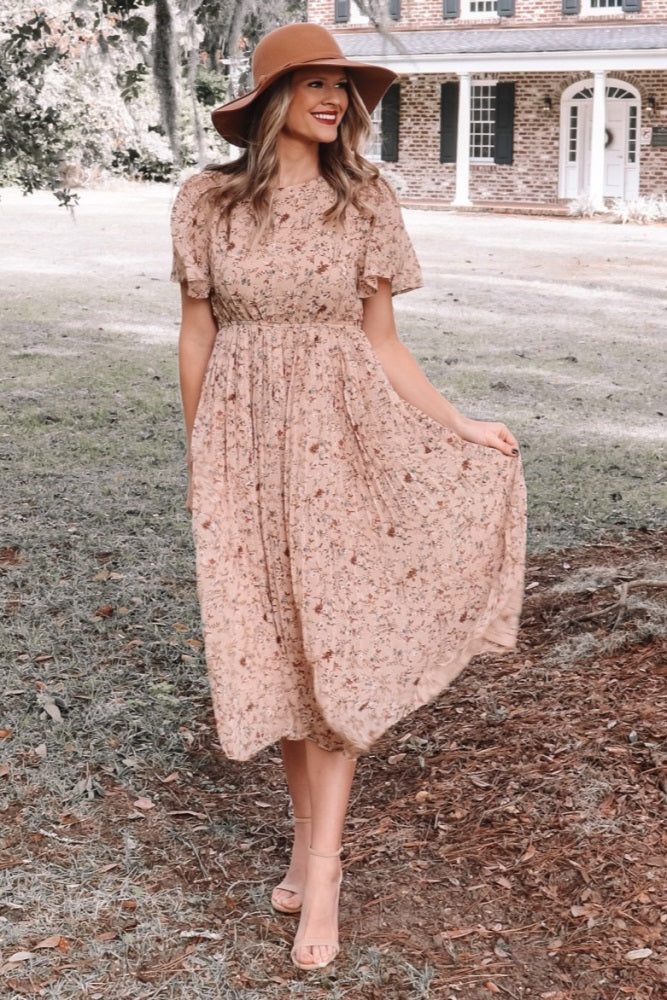 woman wearing a blush pink floral midi dress and a wide-brimmed hat, standing outdoors on a grassy area