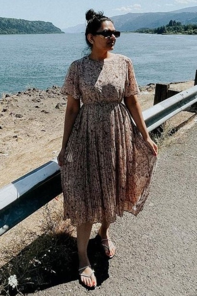woman wearing a floral midi dress with short sleeves and a pleated skirt, standing by a riverbank in sunglasses