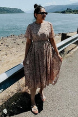 woman wearing a floral midi dress with short sleeves and a pleated skirt, standing by a riverbank in sunglasses