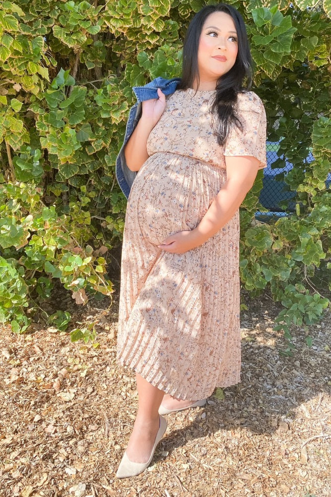 woman wearing a blush floral pleated maternity dress, posing with a baby bump and holding a denim jacket