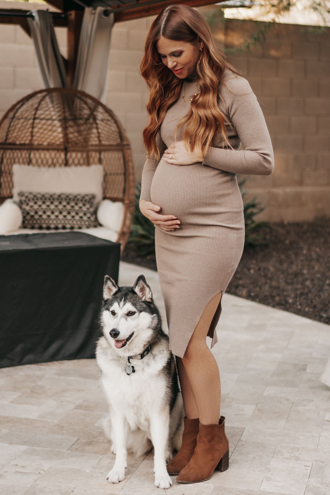 woman wearing a beige ribbed maternity dress with a side slit and holding her baby bump, standing next to a dog