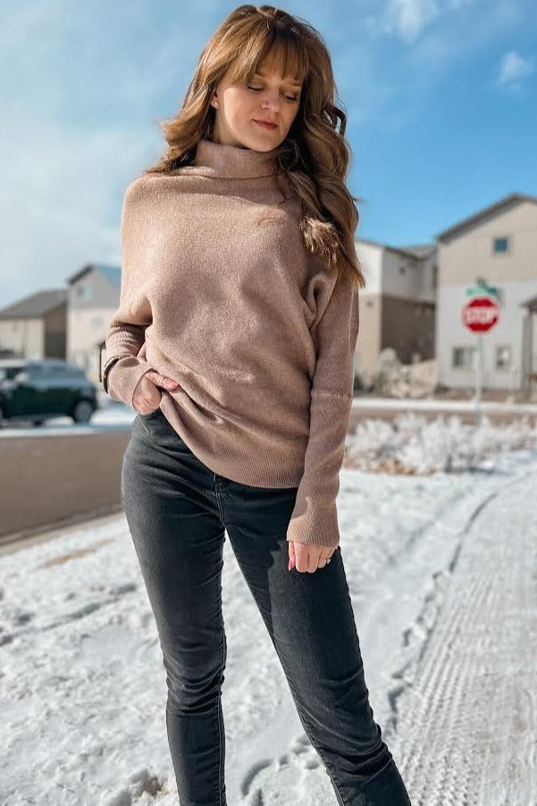 woman wearing a beige turtleneck sweater and black skinny jeans, standing outdoors in a snowy setting
