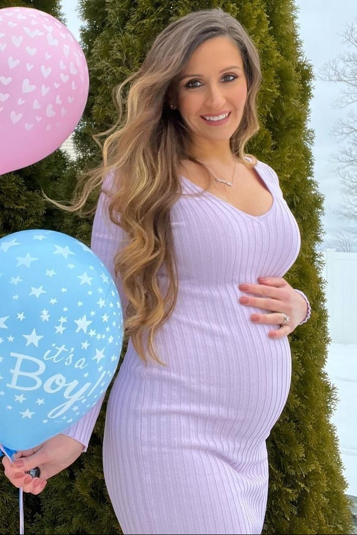 woman wearing a light purple ribbed maternity dress and holding blue and pink balloons near a snowy background with trees