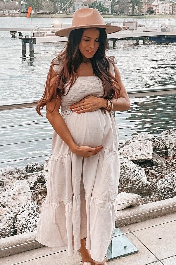 pregnant woman in beige maxi dress and wide brim hat holding her baby bump by a waterfront pier