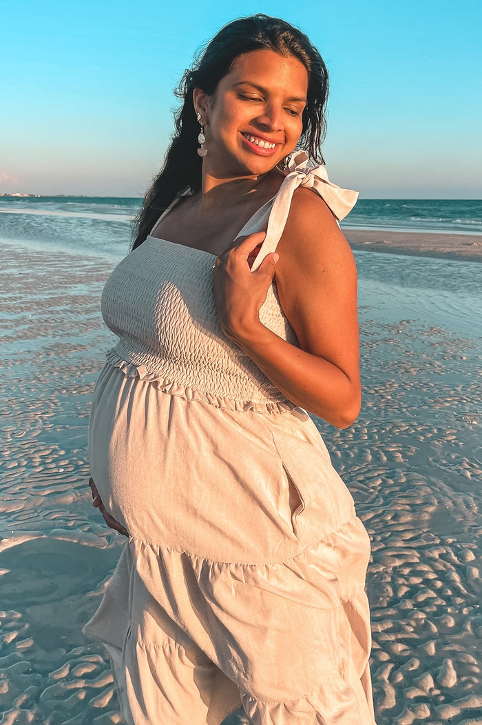 woman wearing a cream-colored maternity dress with a smocked bodice and tiered skirt, showing her baby bump on the beach