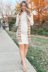 woman wearing a light beige maternity dress with a baby bump and holding a plaid scarf, standing on a sidewalk