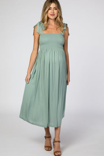 woman wearing a sage green maternity dress with tie shoulder straps and a smocked bodice, featuring a flowing skirt and visible baby bump