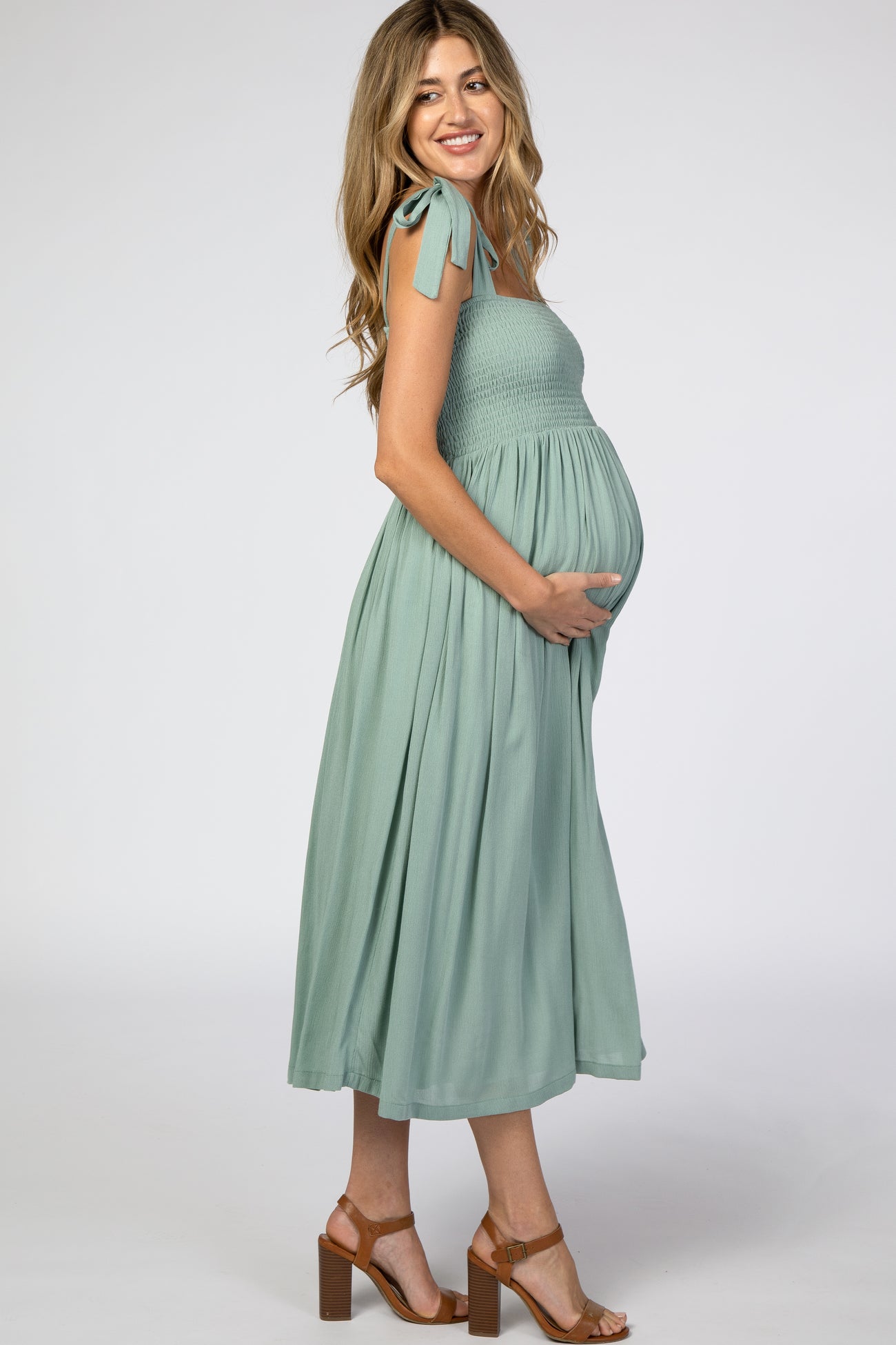 woman wearing a sage green smocked maternity dress with bow tie straps and holding her baby bump