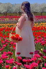 woman wearing a cream maternity dress with ruffle straps, holding a basket of flowers and showing her baby bump in a flower field