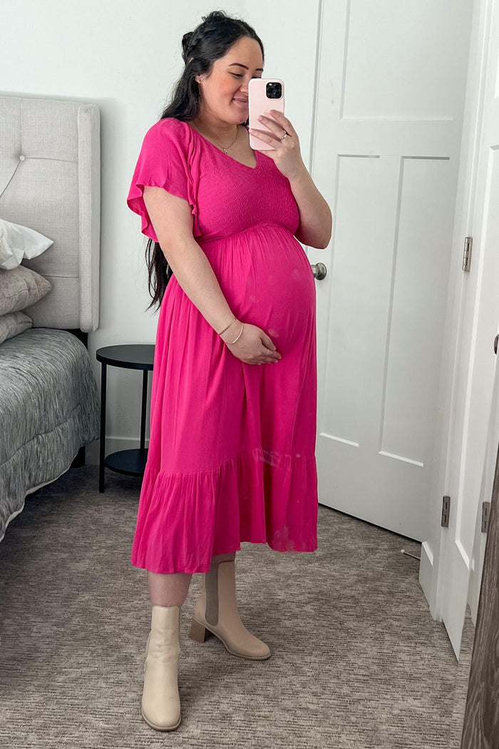 woman wearing a hot pink maternity dress with flutter sleeves, holding her baby bump in a mirror selfie