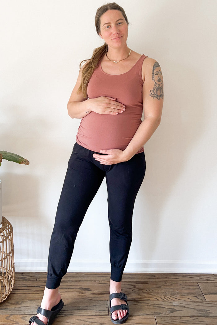 woman wearing a rust-colored maternity tank top and black maternity joggers, holding her baby bump