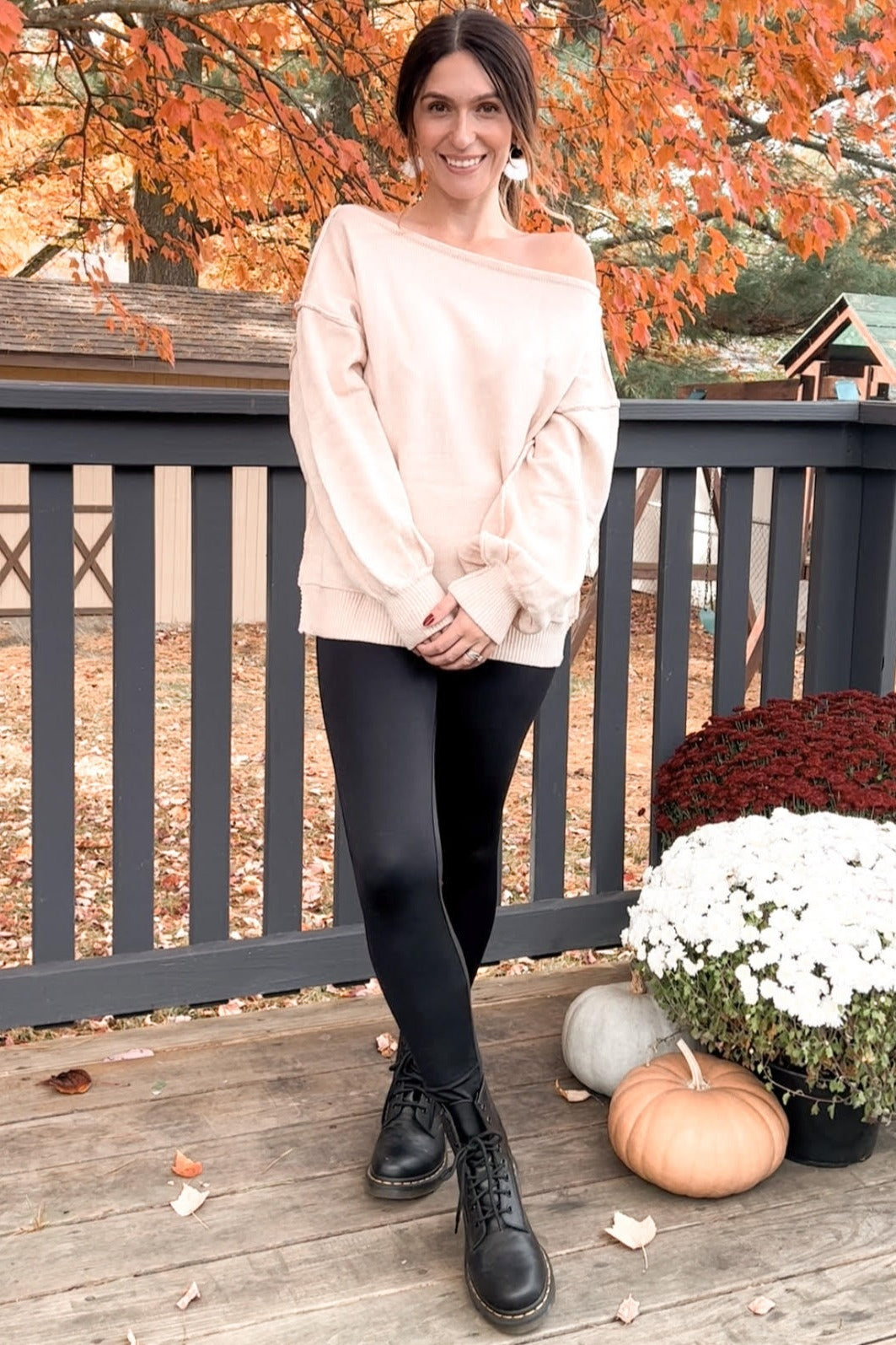 woman wearing a light pink oversized off-the-shoulder sweater, black leggings, and black boots, posing outdoors in autumn setting