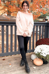 woman wearing a light pink oversized off-the-shoulder sweater, black leggings, and black boots, posing outdoors in autumn setting