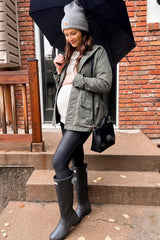 woman wearing a green maternity jacket with a baby bump, black leggings, and gray rain boots, holding an umbrella