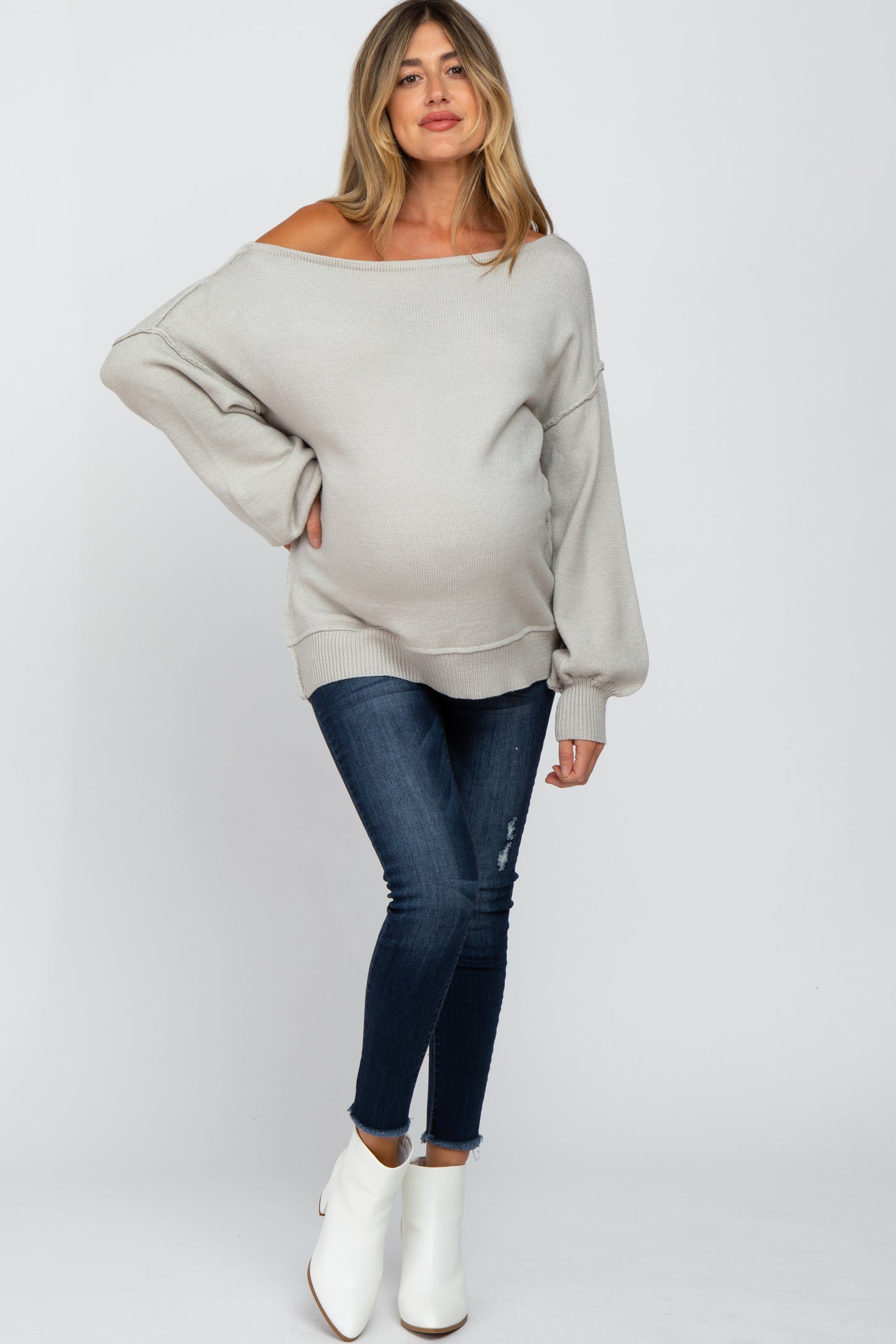 woman wearing a light gray maternity off-shoulder sweater and blue skinny jeans, displaying her baby bump