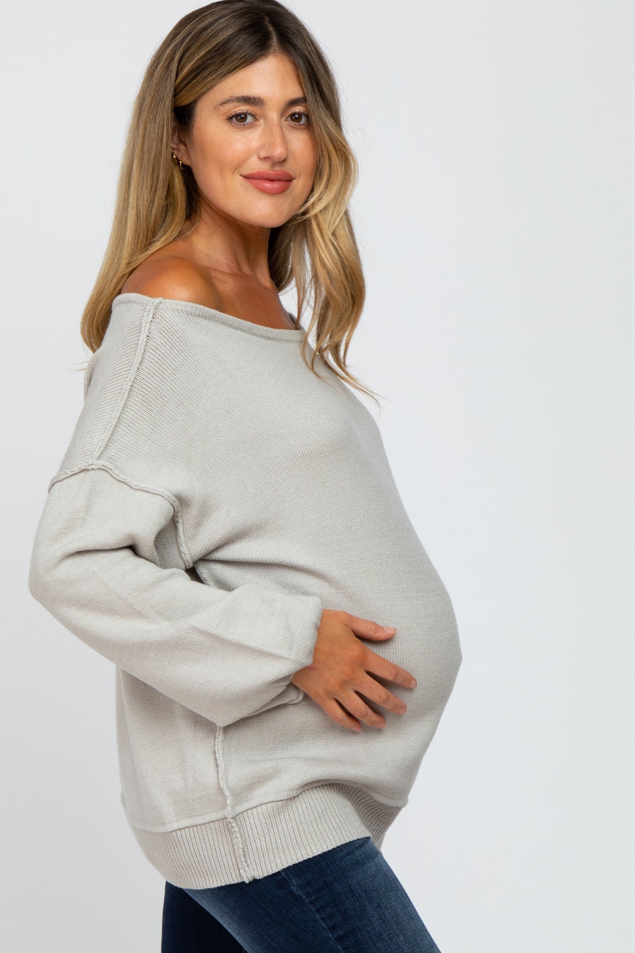 woman wearing a gray off-shoulder maternity sweater and cradling her baby bump