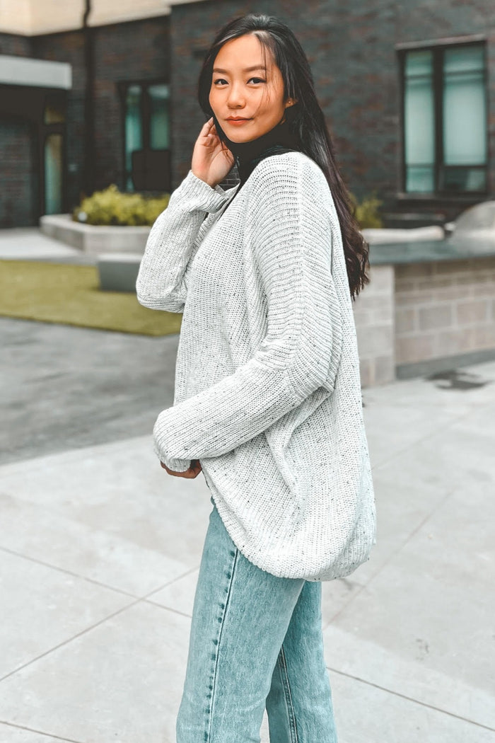 woman wearing a light grey oversized knit sweater and blue high-waisted jeans, posing outside